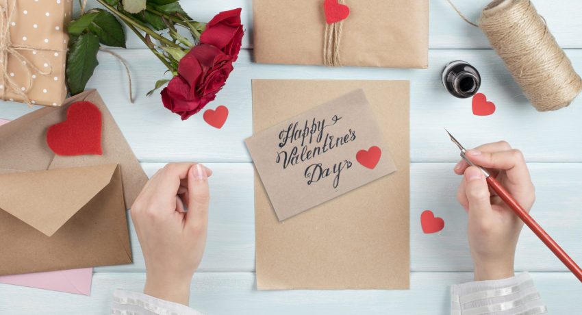 Valentines day theme. Top view of female hands writes greetings. Packed gifts, roses  and envelopes on shabby wooden table. Workplace for preparing handmade decorations.